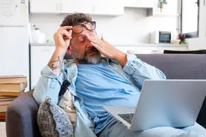 A man is lying on a couch with his hand covering his eyes and a laptop on his lap.