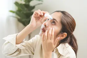A woman is administering eye drops into her eye while holding a bottle in her hand