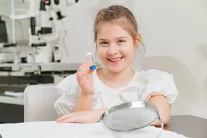 A girl is smiling while holding a tube of medicine and looking at a magnifying glass on a desk in a medical room.