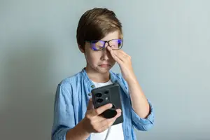 A young boy wearing glasses and a blue shirt is holding a phone with his right hand while covering his eyes with his left hand.