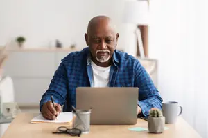 An older man sits at a desk with a laptop, notebook, and pens, possibly working from home.