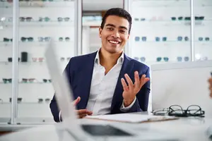 A smiling man in a blue suit and white shirt is sitting at a desk with a laptop and glasses on display in the background.