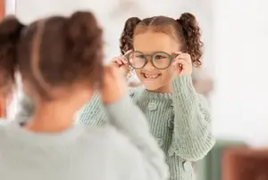 A girl with glasses is smiling at her reflection in a mirror. Another girl is looking at her from the side.