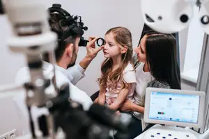 A man is examining a young girl's eyes using an ophthalmoscope, while her mother is watching and the girl is sitting in a chair.