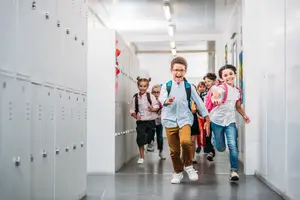 A group of children running down a school hallway, some carrying backpacks.