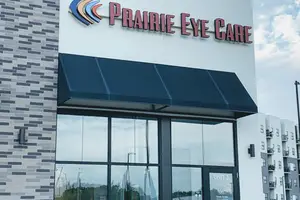 A building with a black awning with the words 'Prairie Eye Care' on it. The building has a brick wall on the left side and glass windows on the right side. The glass windows reflect the surroundings. There is a sign with the text 'UNIT 3' on the door. The sky is cloudy.