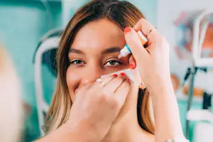 A woman is applying a white cream to her eyebrow area with a stick, while another person is holding her face with their hand. The woman is wearing a ring, bracelet, and necklace. Behind her is a blurry background with a white object, possibly a chair, and a green wall.