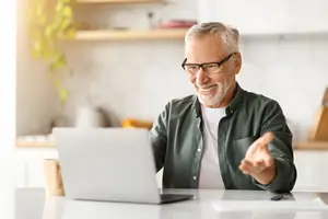 Older man wearing glasses and a green shirt, sitting at a desk with a laptop and a cup, and talking to someone in front of him.