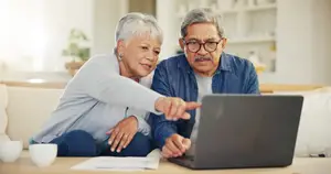 An older couple is sitting on a couch, with the woman pointing at something on a laptop that is placed on a table in front of them.