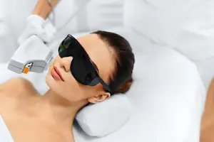 A woman is receiving a laser treatment on her face while lying on a bed with a white pillow and a person wearing white gloves standing behind her.