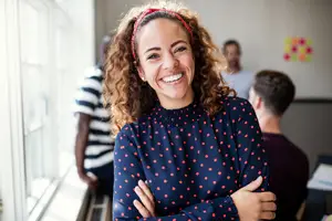 A woman with curly hair wearing a red headband is smiling and posing for a photo while standing in front of a window. Behind her, a man is sitting and looking at something on the right side. On the left side, a person is standing and looking at something. On the wall, there is a yellow and orange sticker.