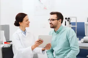 A smiling woman wearing a white lab coat is looking at a tablet with a smiling man wearing glasses in an indoor area