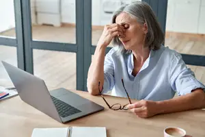 Elderly woman sitting at a desk, holding her forehead with her left hand and resting her chin on her right hand, looking tired