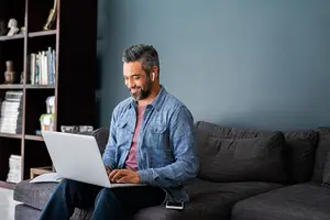 A man is sitting on a couch and smiling while working on his laptop, with a bookshelf in the background.