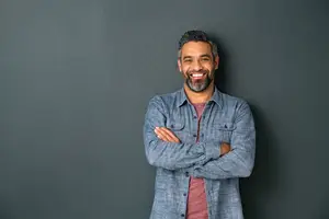 A smiling man with a beard wearing a blue shirt and a red shirt underneath it, standing against a gray wall with his arms crossed