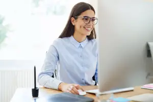 A woman wearing glasses is sitting in front of a desk and using a computer. She is smiling and looking at the camera.