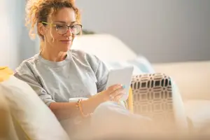 Woman in a gray sweatshirt reading a tablet while sitting on a couch with pillows