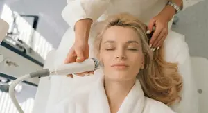 A woman is receiving a facial treatment with a device while lying on a bed, wearing a white robe, and has her eyes closed