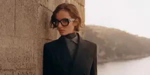 A woman in a black suit and glasses leaning against a wall with a view of the sea in the background
