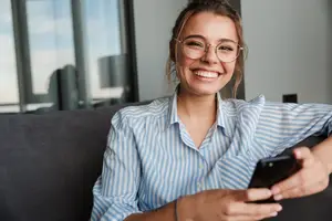 A woman in a blue and white striped shirt sits on a couch and smiles while looking at her phone.