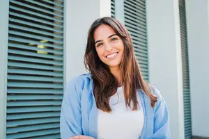 A smiling woman wearing a blue jacket and white shirt stands outside a building with blue shutters
