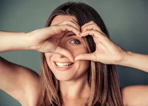 A woman with brown hair and a heart shape made by her hands is posing for a photo with a smile on her face.