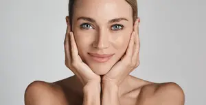 A smiling woman with her hands on her face, wearing a white dress, with blue eyes and brown hair, possibly posing for a photograph.