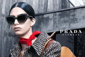 A woman wearing Prada sunglasses with a red scarf and checkered coat stands outside against a gray wall