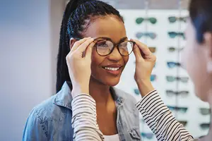 A smiling woman with glasses is being helped by another woman in a shop with shelves of glasses behind them