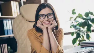 A smiling woman with glasses is sitting in an office chair with her hands on her cheeks.