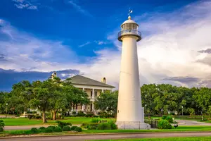 A lighthouse stands tall near a house with a gray roof on a grassy field under a partly cloudy sky