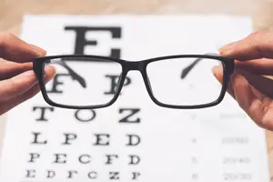 A person is holding a pair of black glasses with a white eye chart in the background.