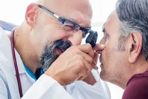 An adult man wearing glasses and a lab coat is examining the eyes of an older man wearing a maroon shirt with an ophthalmoscope.