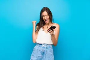 A woman smiling and celebrating while holding a phone and wearing a white sleeveless top and denim shorts against a blue wall