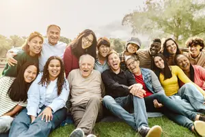 A group of people sitting on the grass, some standing behind them, and smiling, probably posing for a photo in an outdoor area, with trees in the background.