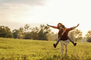A man carrying a woman on his shoulders in a grassy field with trees in the background
