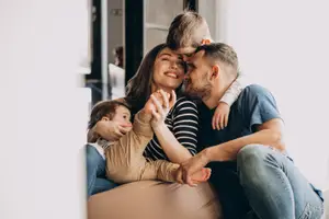 A happy family sitting together in a living room, with a child sitting on the father's lap and another on the mother's lap, all of them smiling and enjoying their time together.