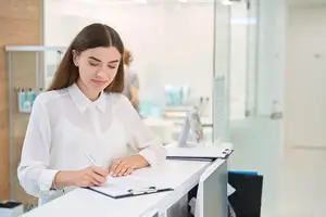 A woman sitting at a desk writing in a notebook while looking down.