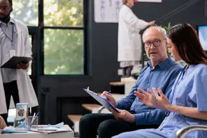 An older man wearing glasses talks to a woman in a blue scrub in a hospital office