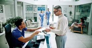 A man is talking to a man in blue scrubs in a hospital. The man in scrubs is sitting on a chair and holding a paper and a pen. The man is holding a paper and a pen and is looking at the man in scrubs. There are other people in the background, including a woman sitting on a chair and a man in blue scrubs walking.