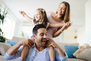 A man sitting on a couch with a girl sitting on his shoulder and a woman standing behind him smiling
