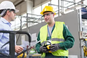 Two men in protective gear and hard hats stand in a factory setting, one holding a paper and the other holding a pair of ear protection