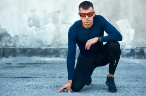 A man in a blue long-sleeved shirt and black pants is kneeling on the ground in front of a wall, possibly preparing for a workout or taking a break.