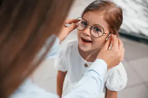 A young girl is wearing glasses and is smiling while a woman adjusts her glasses.