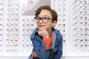 a smiling boy with glasses sits in front of a wall of glasses in an optical store