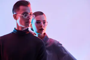 An adult man and woman with glasses on their faces pose for a picture with a pink background