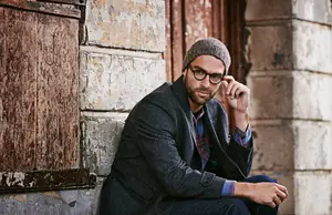 A man with a beanie and glasses is sitting on a brick wall and posing for a photo.
