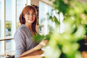 A woman in glasses is standing inside a coffee shop and smiling at the camera