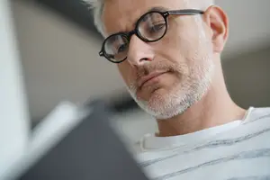 An older man with white hair and a beard is reading a book and wearing glasses
