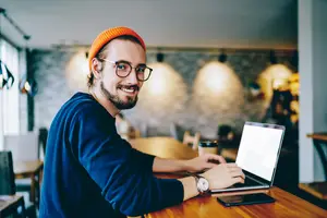 Man in blue long sleeve shirt and orange beanie sitting at a table in a cafe working on his laptop.
